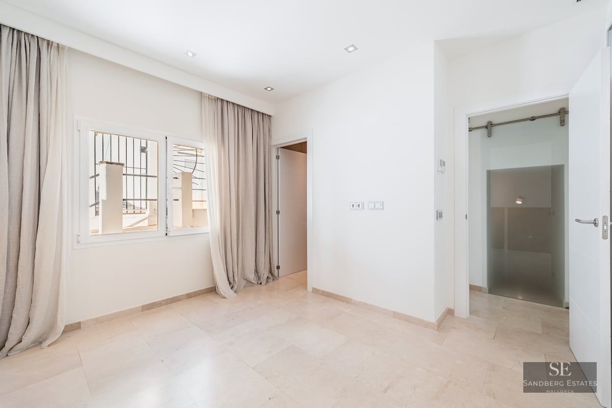 Bright empty room with white walls, cream marble floor, window with floor-length beige curtains, and open doorways.