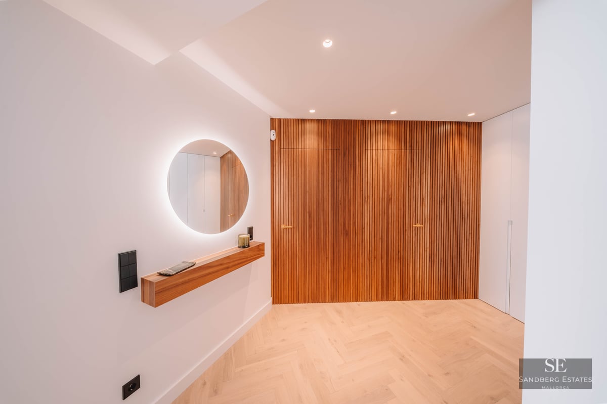 Minimalist hallway featuring a backlit circular mirror, wooden floating shelf, and vertical wood slat wall.