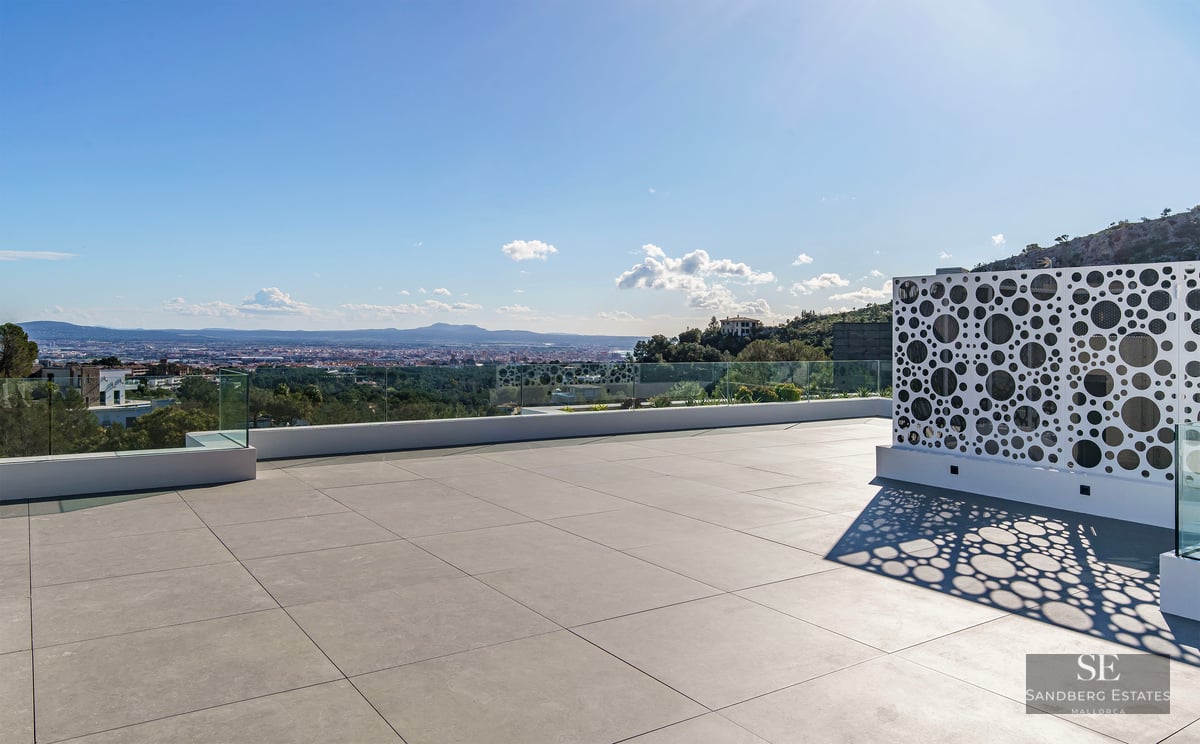 A spacious modern rooftop terrace with grey tiles, glass railings, and a white perforated screen overlooking a city.