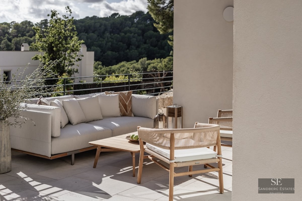Modern outdoor seating area on a terrace with a white sofa, wooden chairs, and views of a lush green forest.