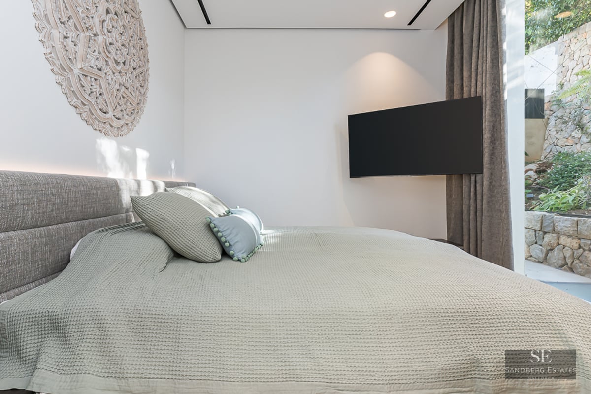 Modern bedroom featuring a grey bed, wall-mounted TV, and a large circular carved wooden ornament on a white wall.