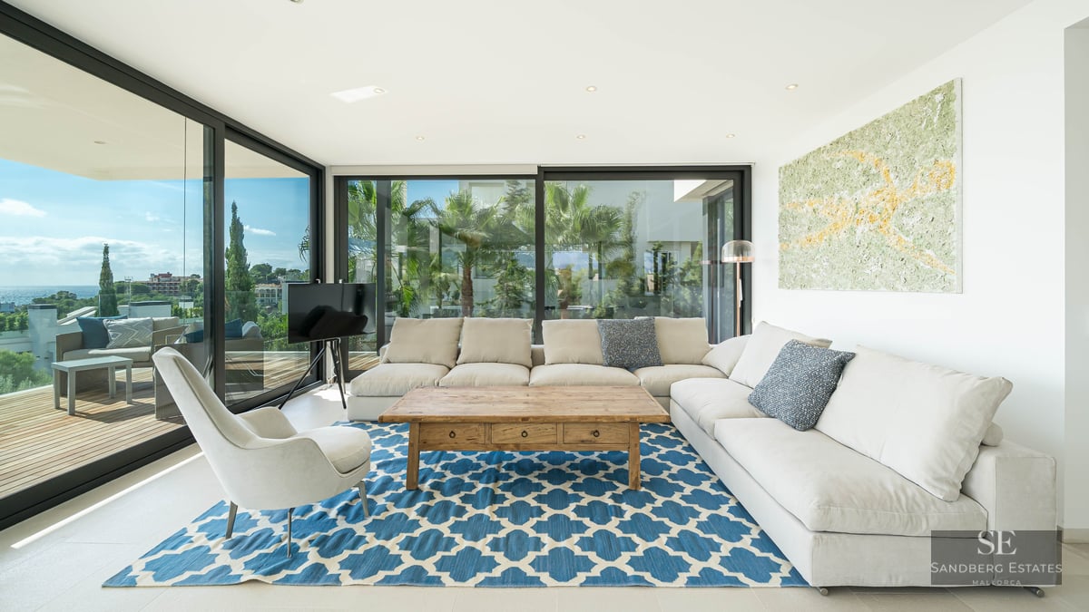 Modern living room with white L-shaped sofa, blue patterned rug, and floor-to-ceiling windows showing a coastal view.