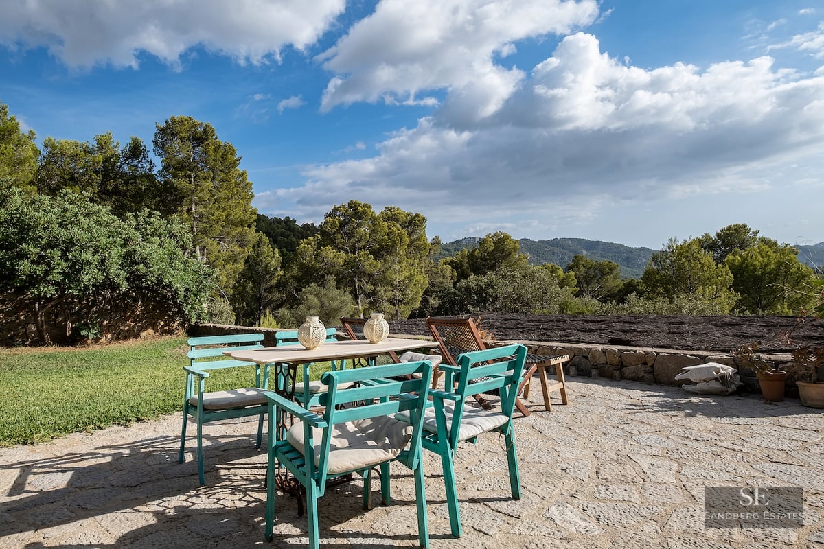 Stone terrace with a white table and turquoise chairs surrounded by forest and mountains under a blue sky.