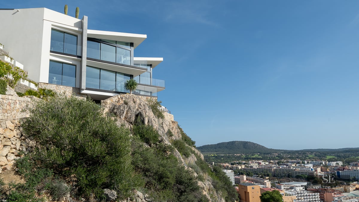 Modern white villa with large glass windows perched on a rocky cliff overlooking a city landscape under a blue sky.