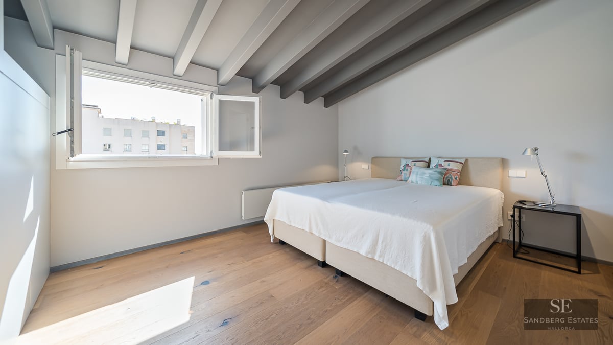 Bright bedroom featuring a double bed, wooden flooring, grey ceiling beams, and a large window.