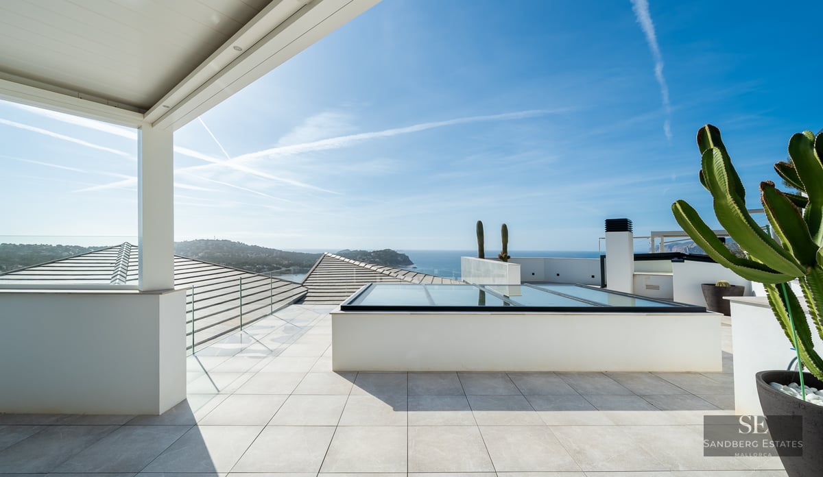 Bright white rooftop terrace with glass railings overlooking the sea and sky with large cacti in pots.