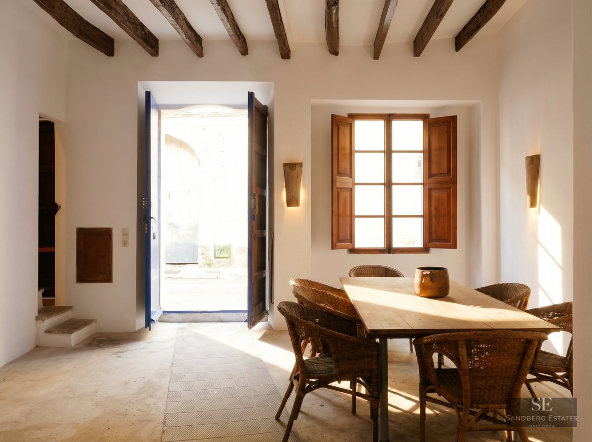A rustic dining room with a large wooden table, wicker chairs, exposed ceiling beams, and a sunlit open doorway.