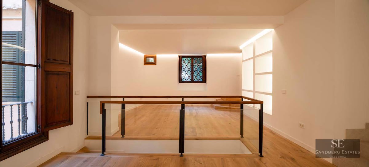 A modern indoor landing with light oak flooring, glass railings, and warm cove lighting against white walls.