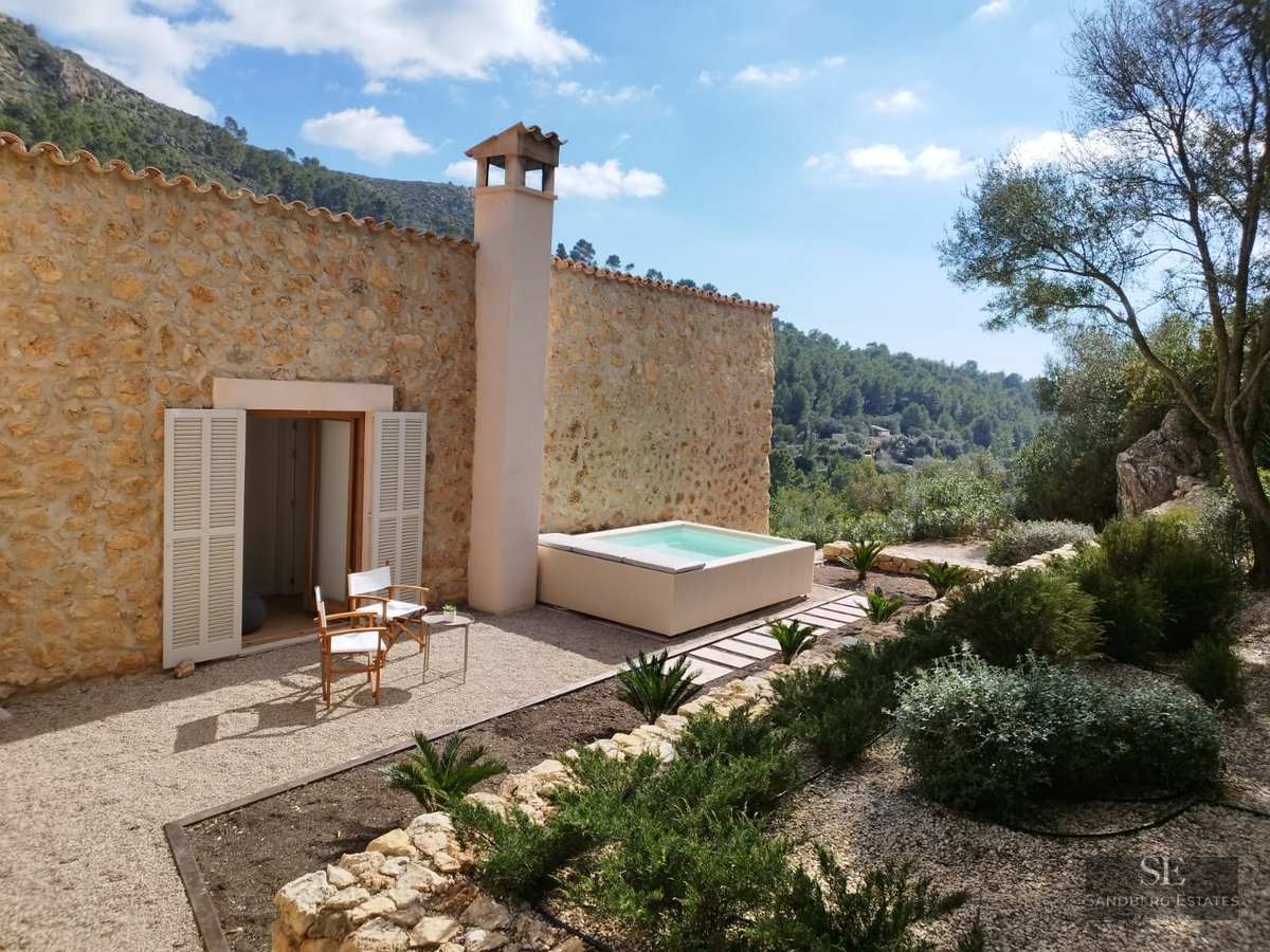 Stone house featuring an outdoor jacuzzi, wooden chairs on gravel, and mountain views under a clear sky.