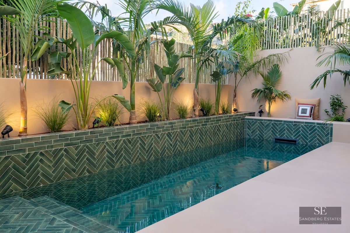 Piscine de luxe avec carreaux verts en chevrons, palmiers et murs beiges avec un éclairage chaleureux.