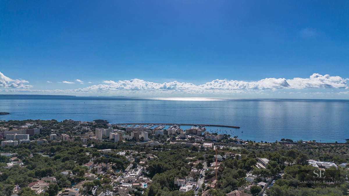 Vue aérienne d'une ville côtière avec marina, arbres verts et mer Méditerranée sous un ciel bleu clair.