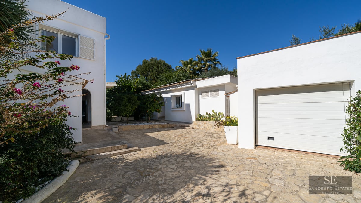 White Mediterranean villa with a stone-paved driveway, garage, and lush greenery under a clear blue sky.
