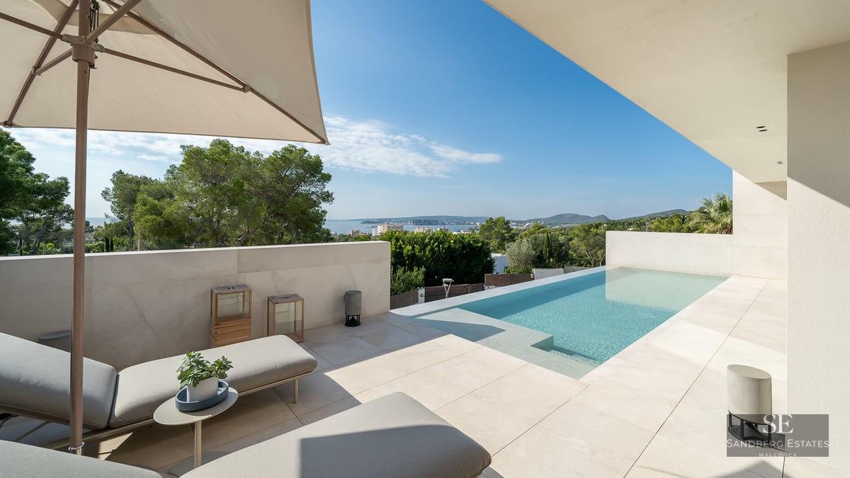 Sun loungers on a modern beige stone terrace next to a swimming pool overlooking the sea and green landscape under a clear sky.