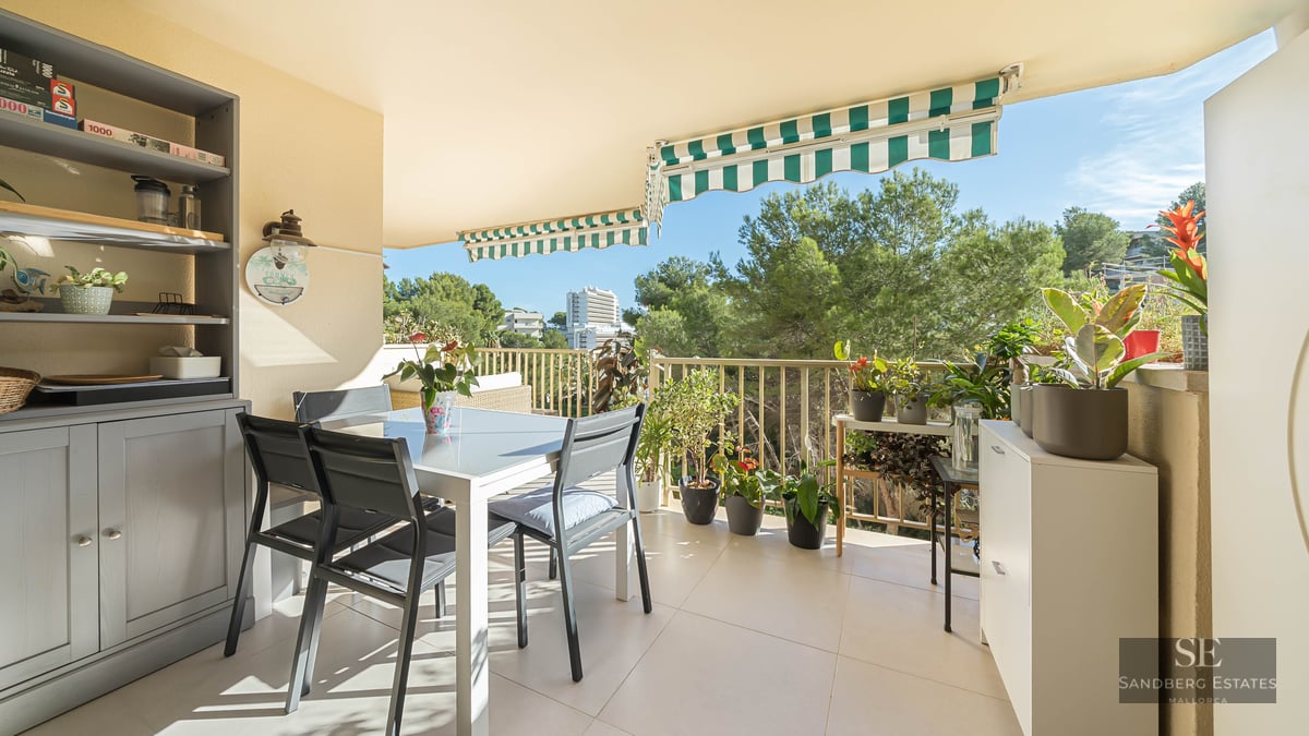 Un balcon ensoleillé avec une table à manger blanche, des chaises sombres, de nombreuses plantes en pot et des stores rayés.