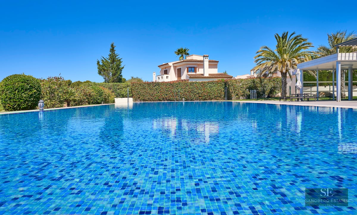 Large blue mosaic pool under a clear sky, with a Mediterranean villa and palm trees in the background.