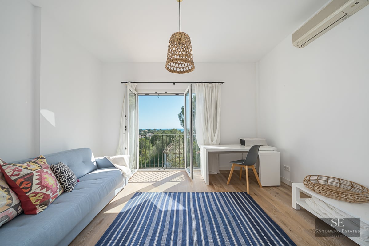 Pièce lumineuse avec canapé bleu, bureau blanc et portes-fenêtres ouvertes sur un paysage verdoyant et la mer.