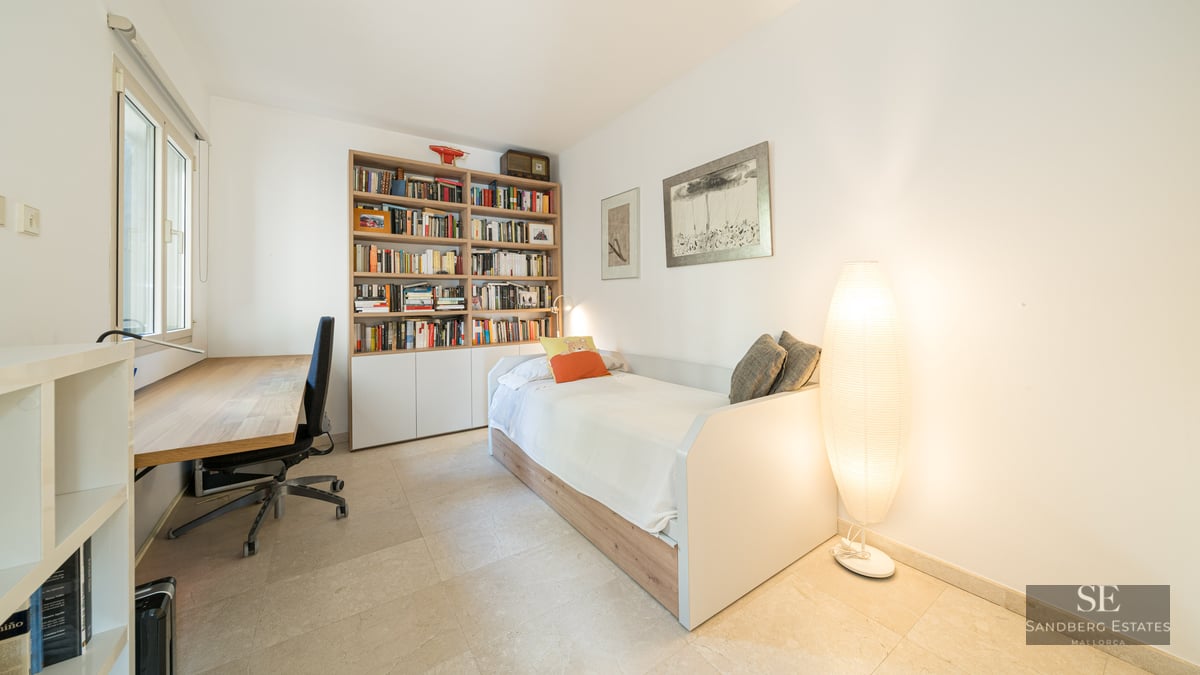Bright bedroom featuring a single bed, a large wooden bookshelf, a desk with a chair, and a glowing floor lamp.