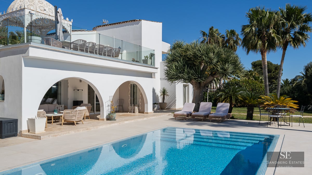 White arched villa overlooking a turquoise swimming pool with sun loungers and palm trees under a clear blue sky.