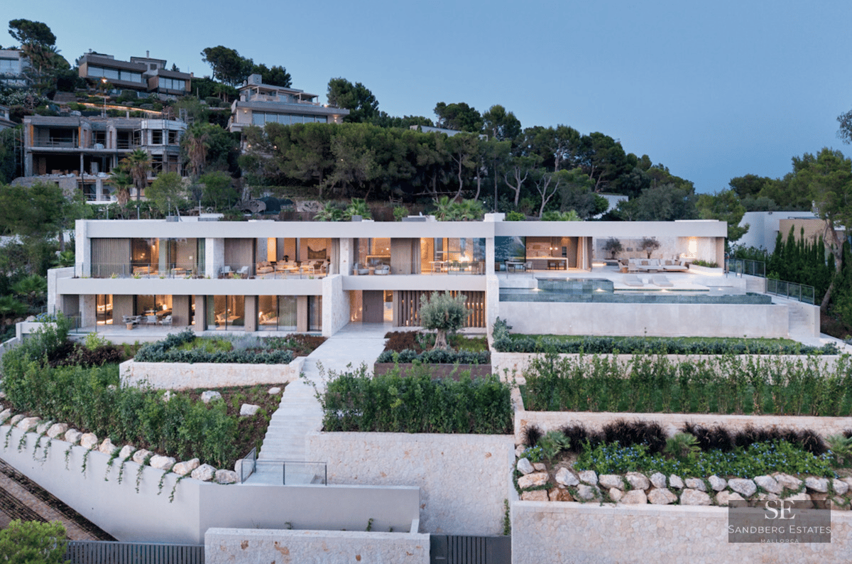 Modern two-story villa with infinity pool, stone terraced gardens, and illuminated glass facade at dusk.