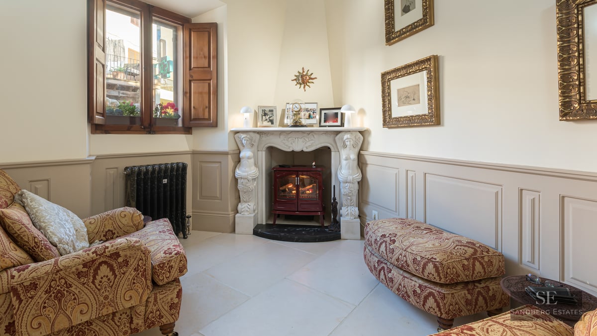 A cozy sitting area featuring an ornate white fireplace with statues, a red stove, and damask-patterned armchairs.