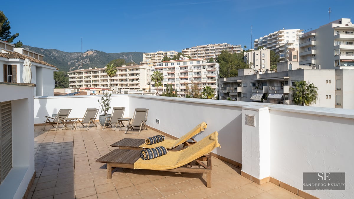 Spacious rooftop terrace featuring wooden sun loungers, tiled flooring, and views of urban buildings and mountains.