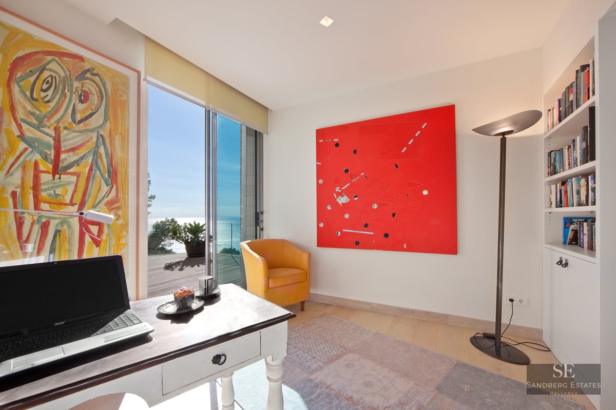 Bright home office with a desk, yellow armchair, abstract art, and sliding doors opening to a sea-view terrace.