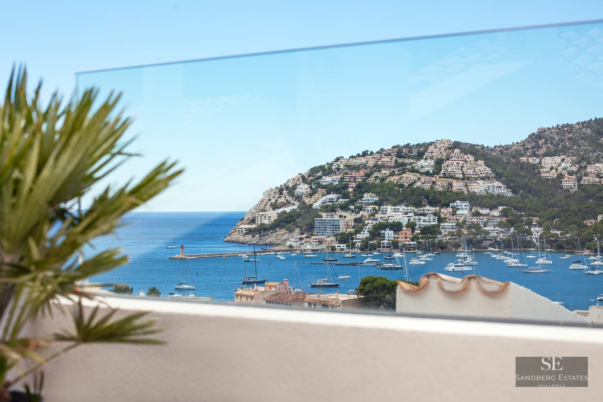 View through a glass balcony railing of a Mediterranean harbor filled with boats and a hillside town.