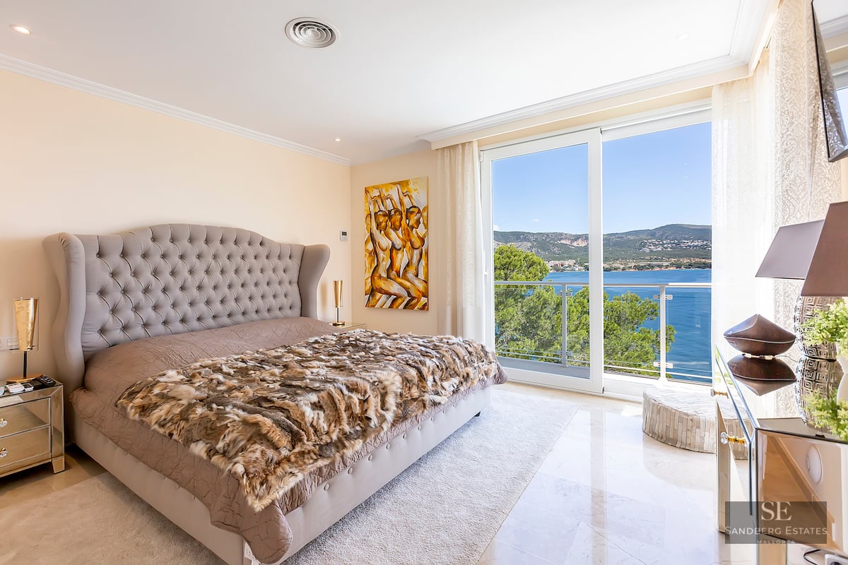 Luxurious bedroom featuring a large tufted bed, marble floors, and sliding glass doors revealing a panoramic sea view.