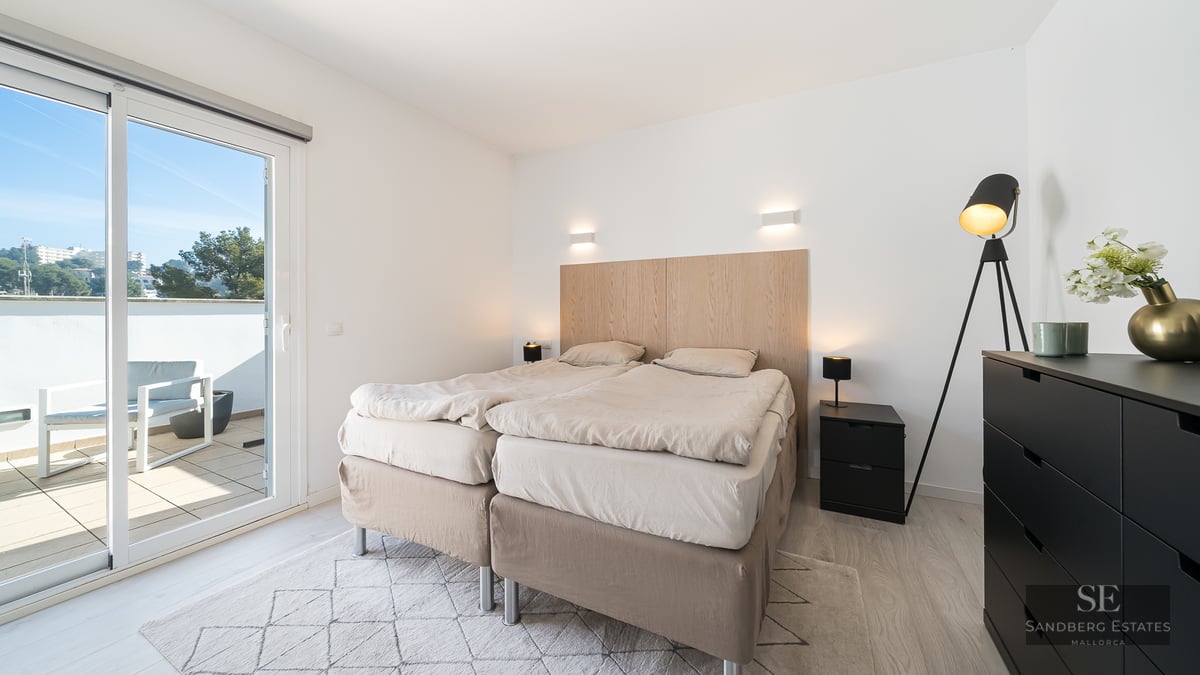 Bright bedroom featuring a double bed, light wood headboard, and a sliding glass door opening to a sunny balcony.