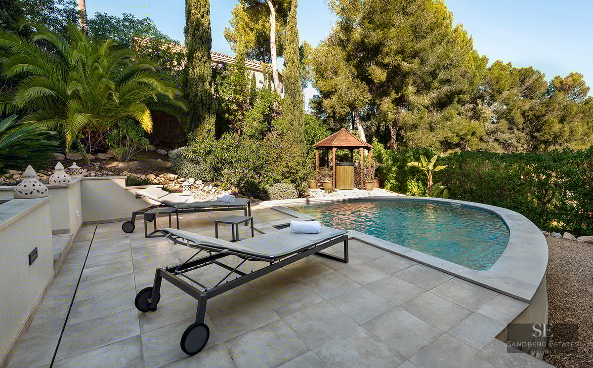 Sun loungers on a stone terrace beside a curved swimming pool surrounded by palm trees and lush Mediterranean greenery.