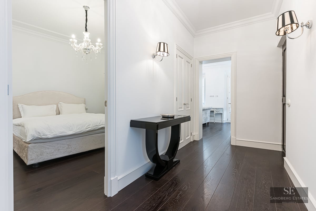Bright hallway with dark wood floors, black console table, and view into a bedroom with a crystal chandelier.