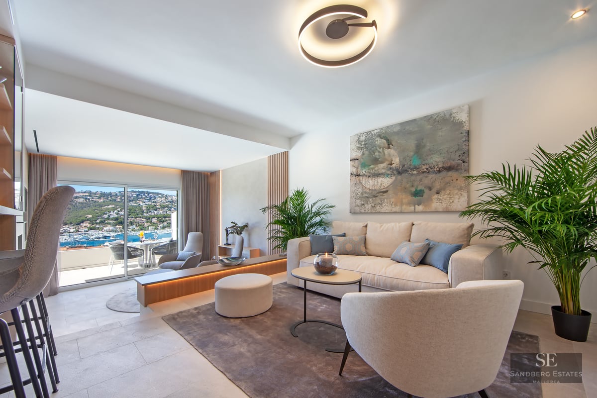 Bright modern living room with cream furniture, large windows overlooking the sea, and lush indoor plants.