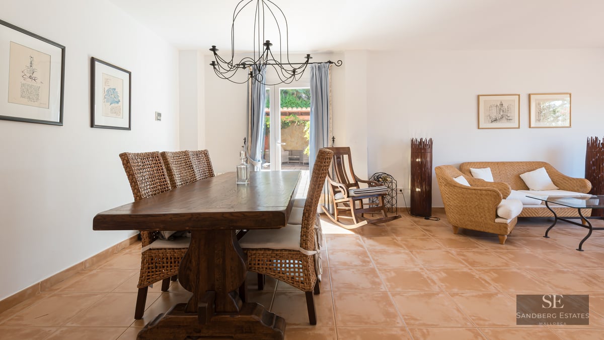 Spacious dining area with a solid wood table and wicker chairs on terracotta tiles, leading to a sunlit lounge area.
