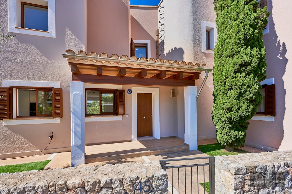 Entrance of a beige villa with a wooden door, stone wall, and a tall cypress tree under a bright blue sky.