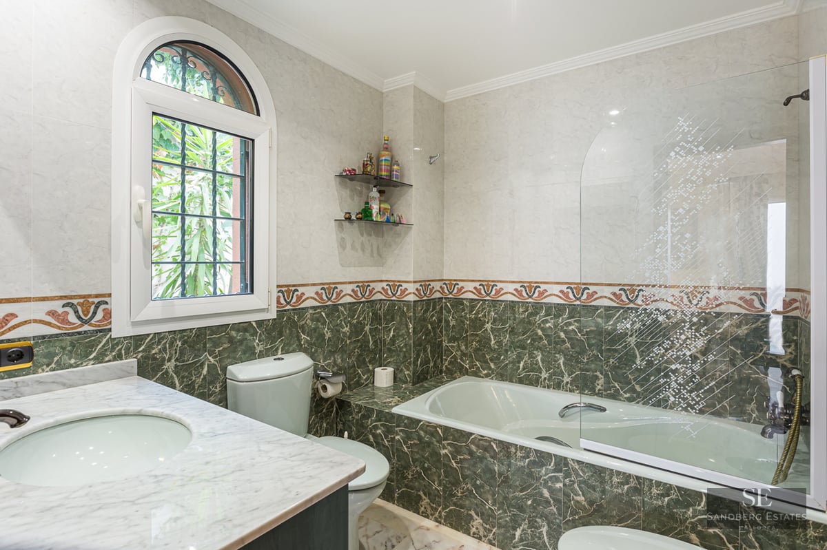 A bathroom featuring green and white marble tiling, a bathtub with a glass screen, and a bright arched window.