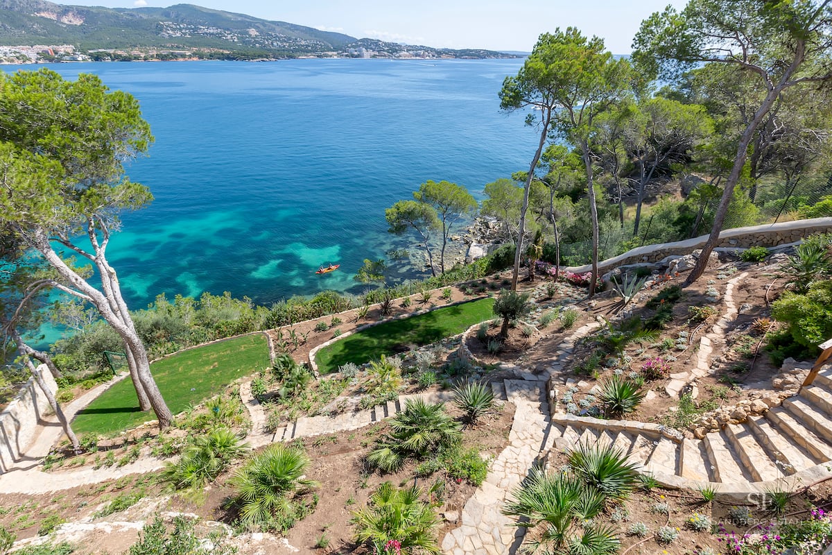 Terraced garden with stone steps and pine trees overlooking a turquoise sea and mountainous coastline.