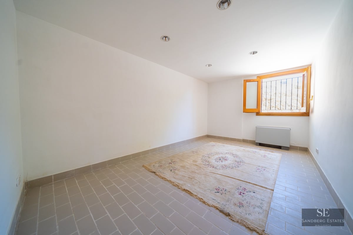 An empty room with light grey floor tiles, white walls, a patterned rug, and a small wooden window.