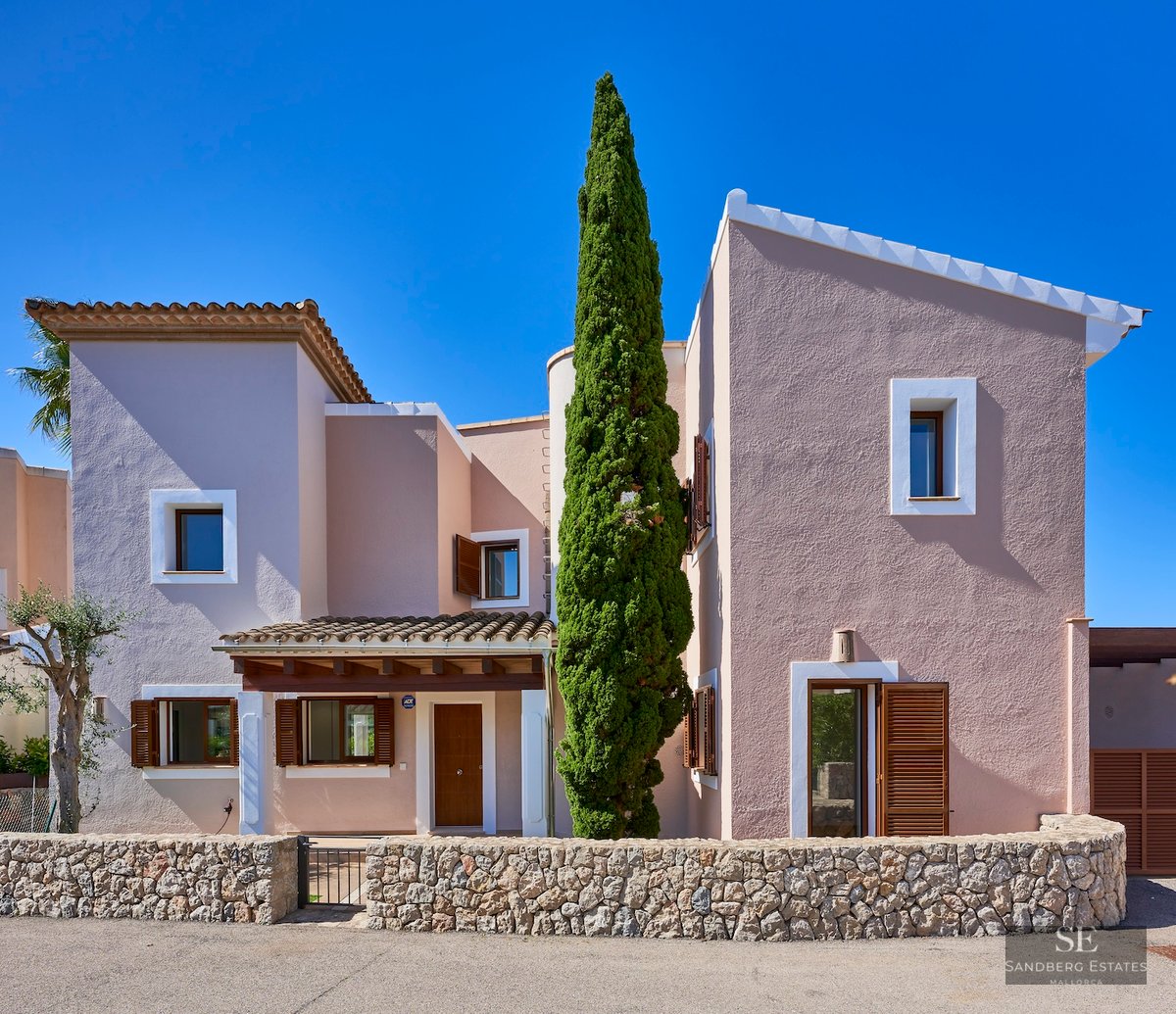 Front view of a pink two-story Mediterranean villa with wooden shutters, a stone wall, and a tall cypress tree.