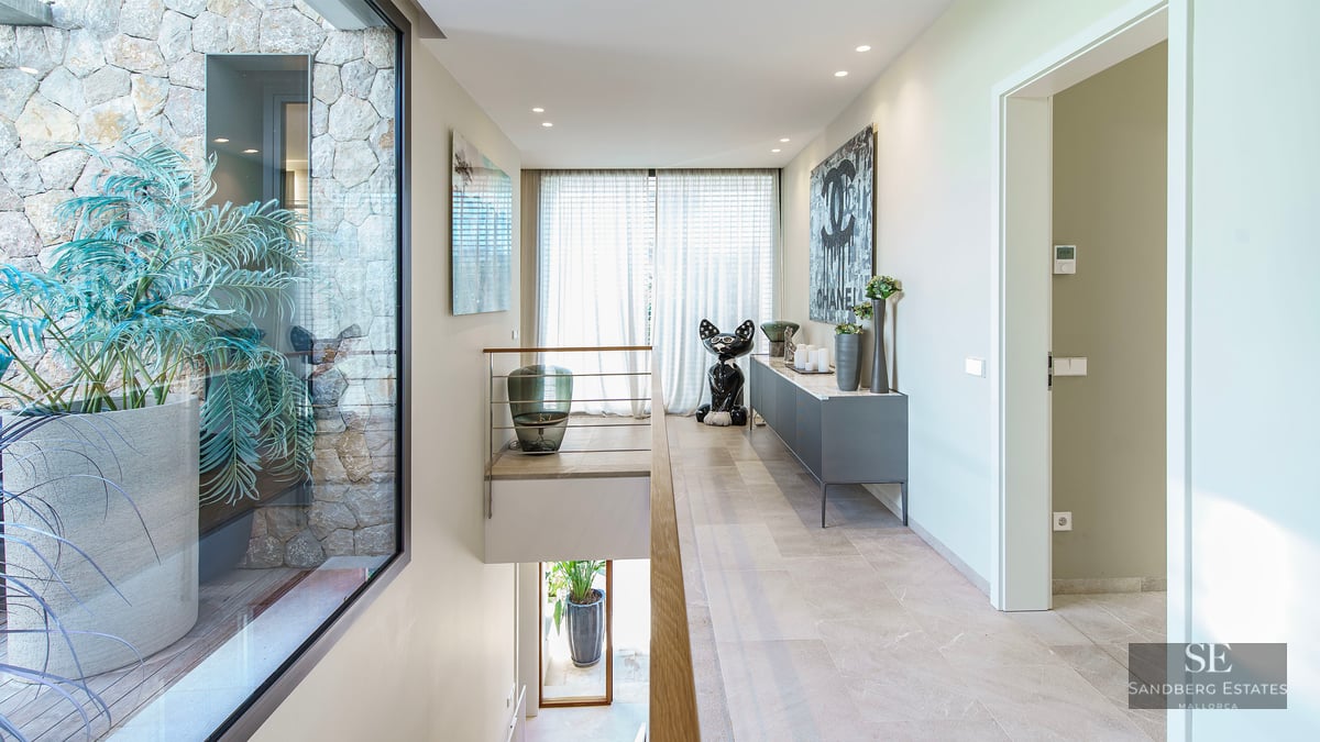 Couloir lumineux, sol en pierre, art moderne et fenêtre donnant sur une cour intérieure avec des plantes.