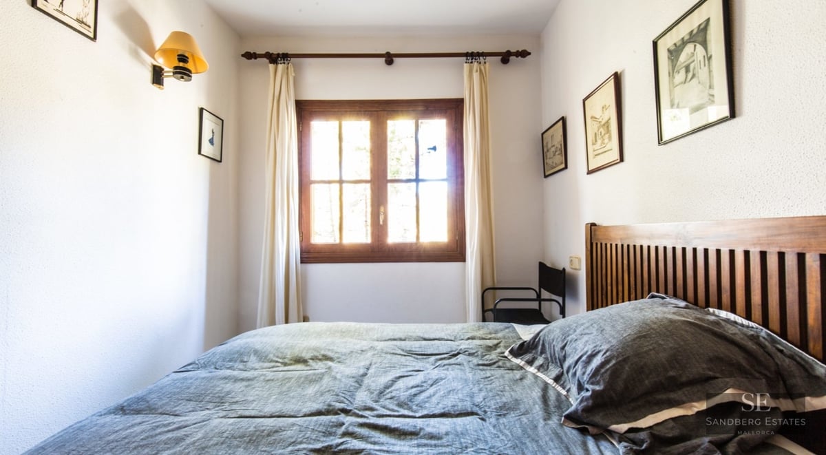 A bedroom with a wooden bed, grey bedding, and a window with bright sunlight coming through white curtains.