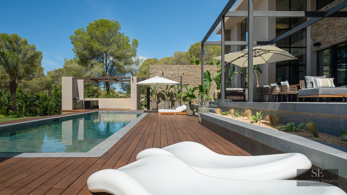 Luxury outdoor swimming pool with wooden deck, white designer sun loungers, and stone walls under a clear blue sky.
