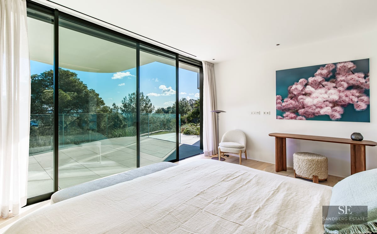 Bright bedroom featuring a white bed, floor-to-ceiling glass doors, and a large terrace overlooking trees.