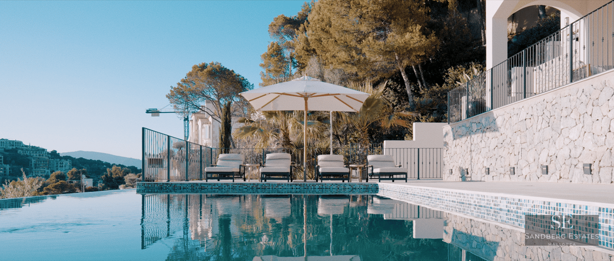 Infinity swimming pool with four sunbeds under a white umbrella, surrounded by stone walls and Mediterranean greenery.
