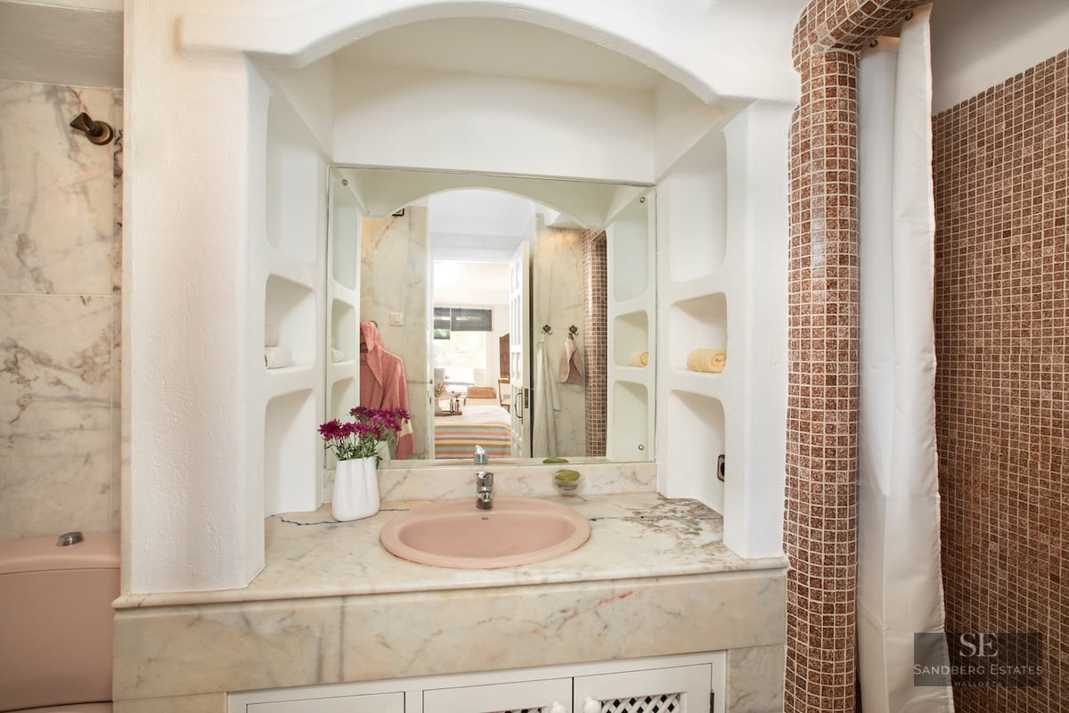 Bathroom vanity with pink sink, marble countertop, arched mirror, and brown mosaic tile pillar.