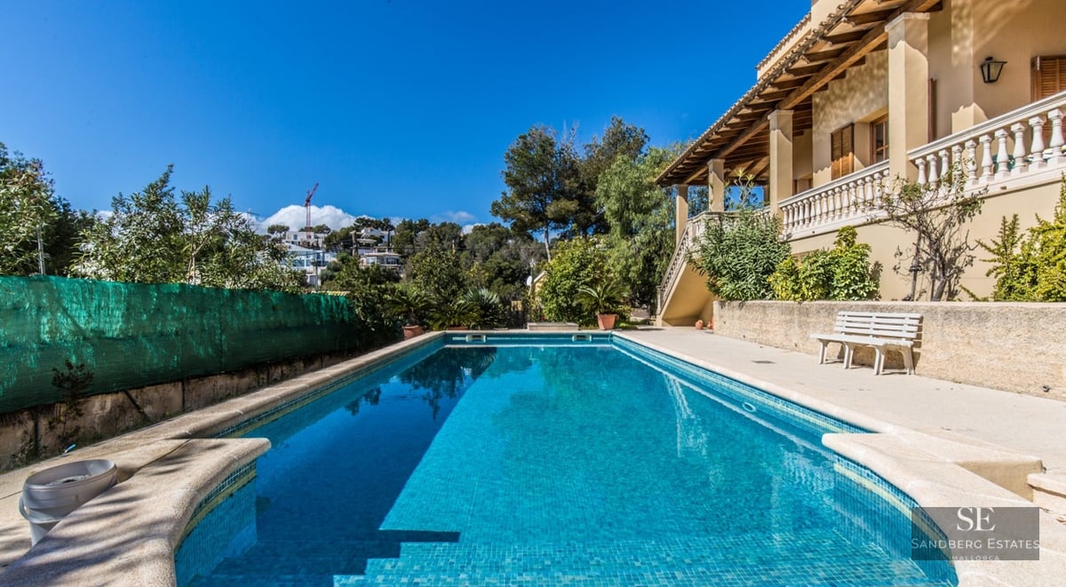 Large rectangular swimming pool with turquoise water next to a Mediterranean villa with a sunny terrace and blue sky.