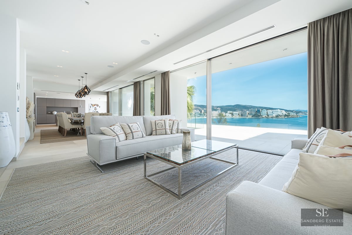 Modern living room with grey sofas and large glass doors opening to a panoramic view of the sea and coastline.