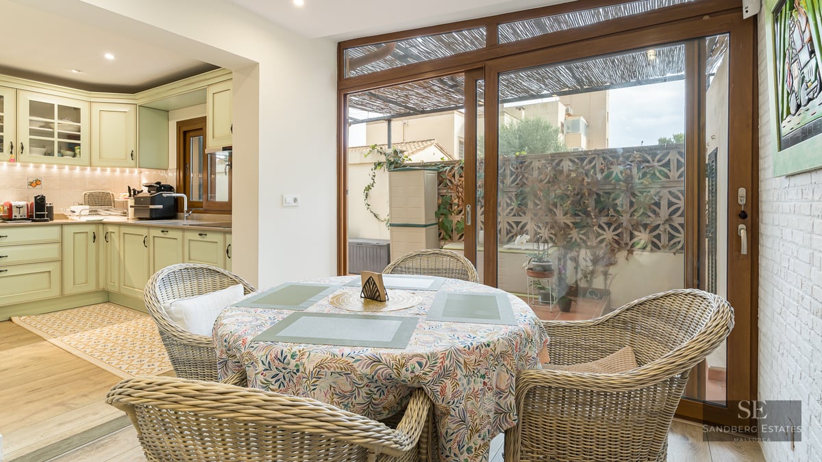 Dining table with wicker chairs next to a sage green kitchen and large sliding glass doors.