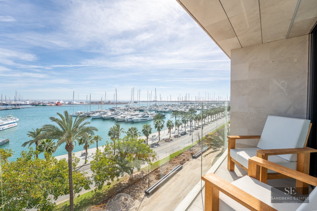 Modern balcony with wooden chairs and glass railing overlooking a marina full of yachts and palm trees.