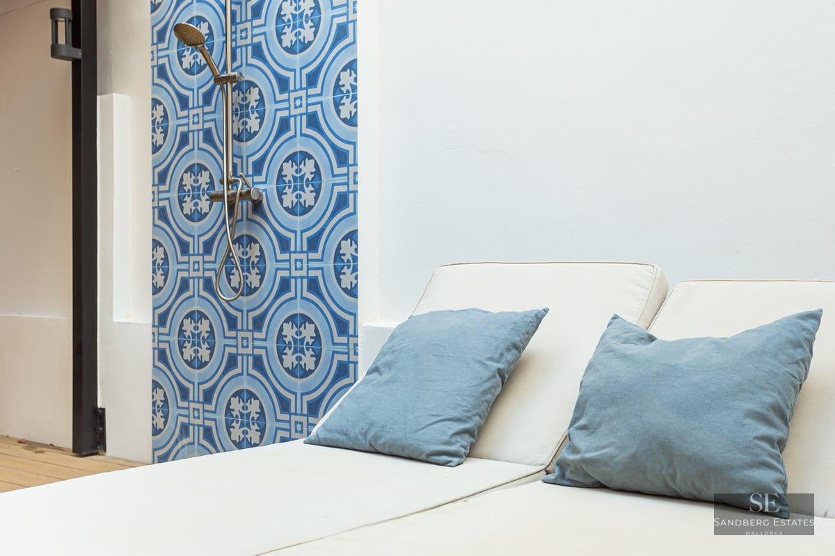 Sun lounger with blue pillows next to a wall with blue patterned tiles and a modern outdoor shower head.