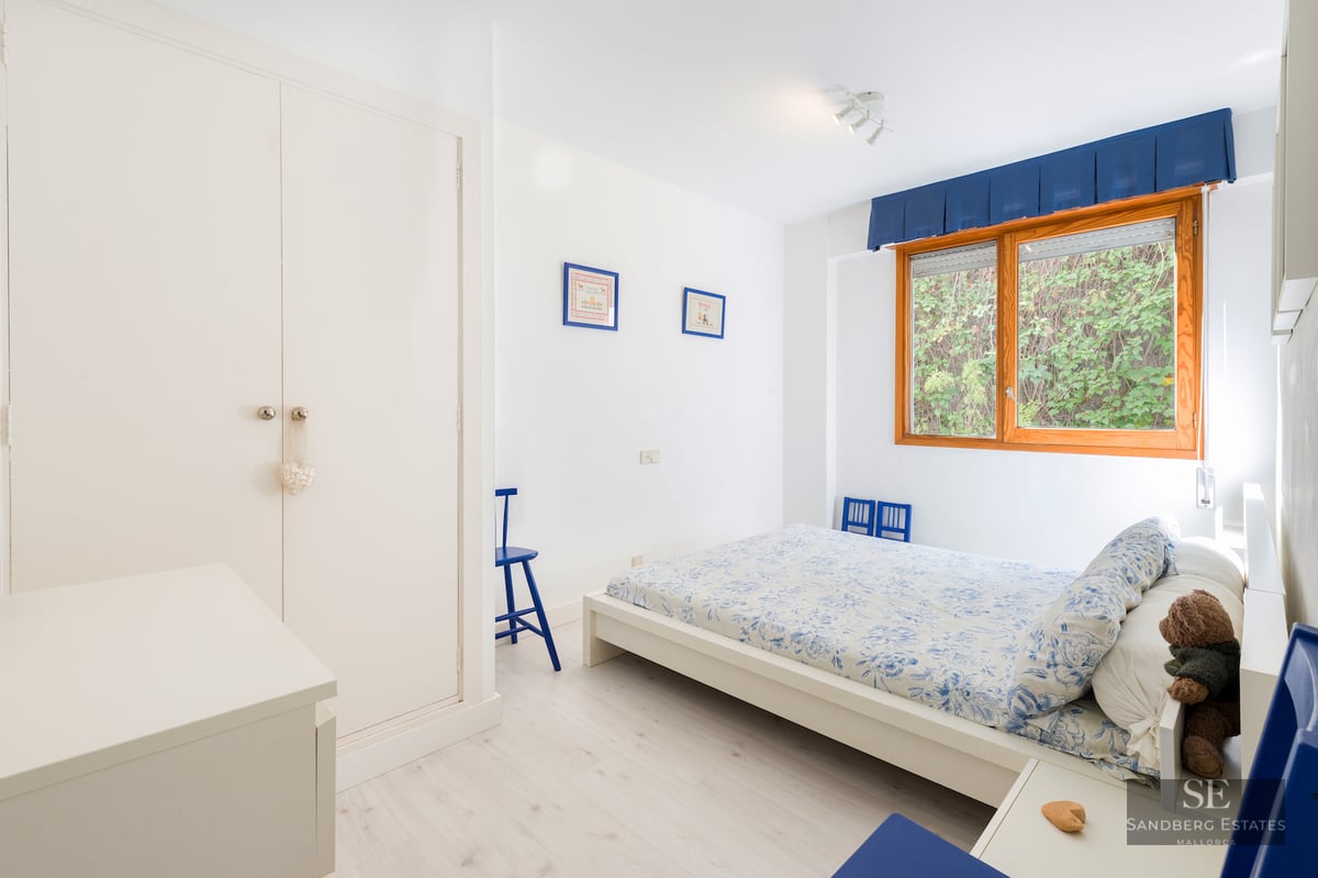 Bright white bedroom featuring a wooden-framed window, built-in wardrobe, and blue and white floral bedding.